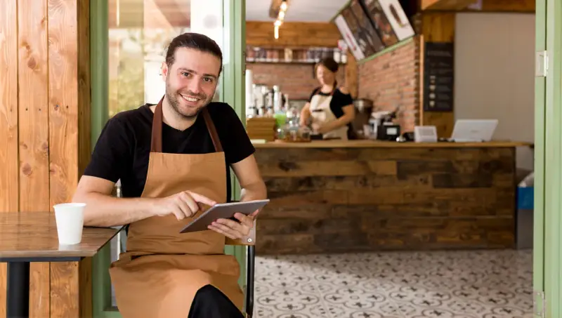 Hombre sonriente con delantal usa tablet en su cafetería, gestionando pedidos con un sistema erp restaurantes.