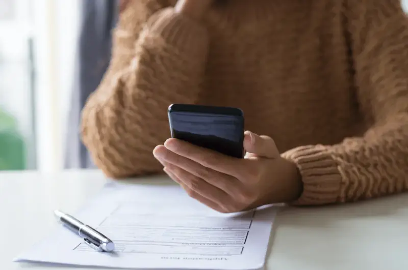 Persona revisa su teléfono móvil, modelo 035, mientras tiene un bolígrafo y un formulario de solicitud sobre la mesa.